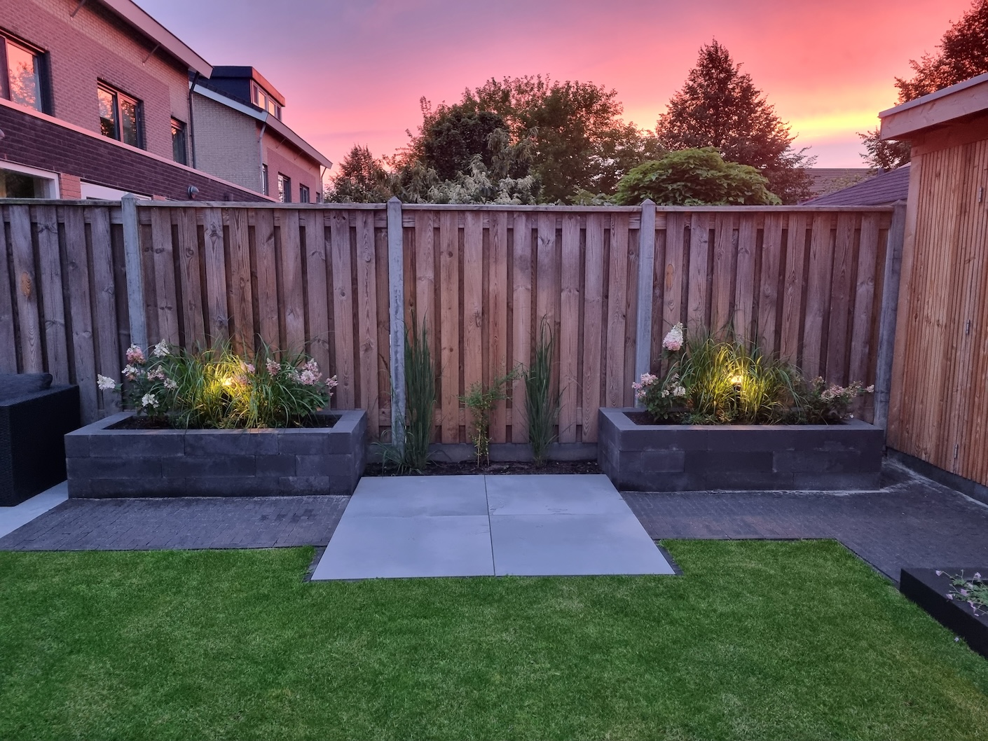 Overzicht van de moderne tuin met een door Snijers Tuinen geplaatste pergola, zitplaatsen, en een goed onderhouden gazon bij zonsondergang. Tuinrenovatie in Lelystad