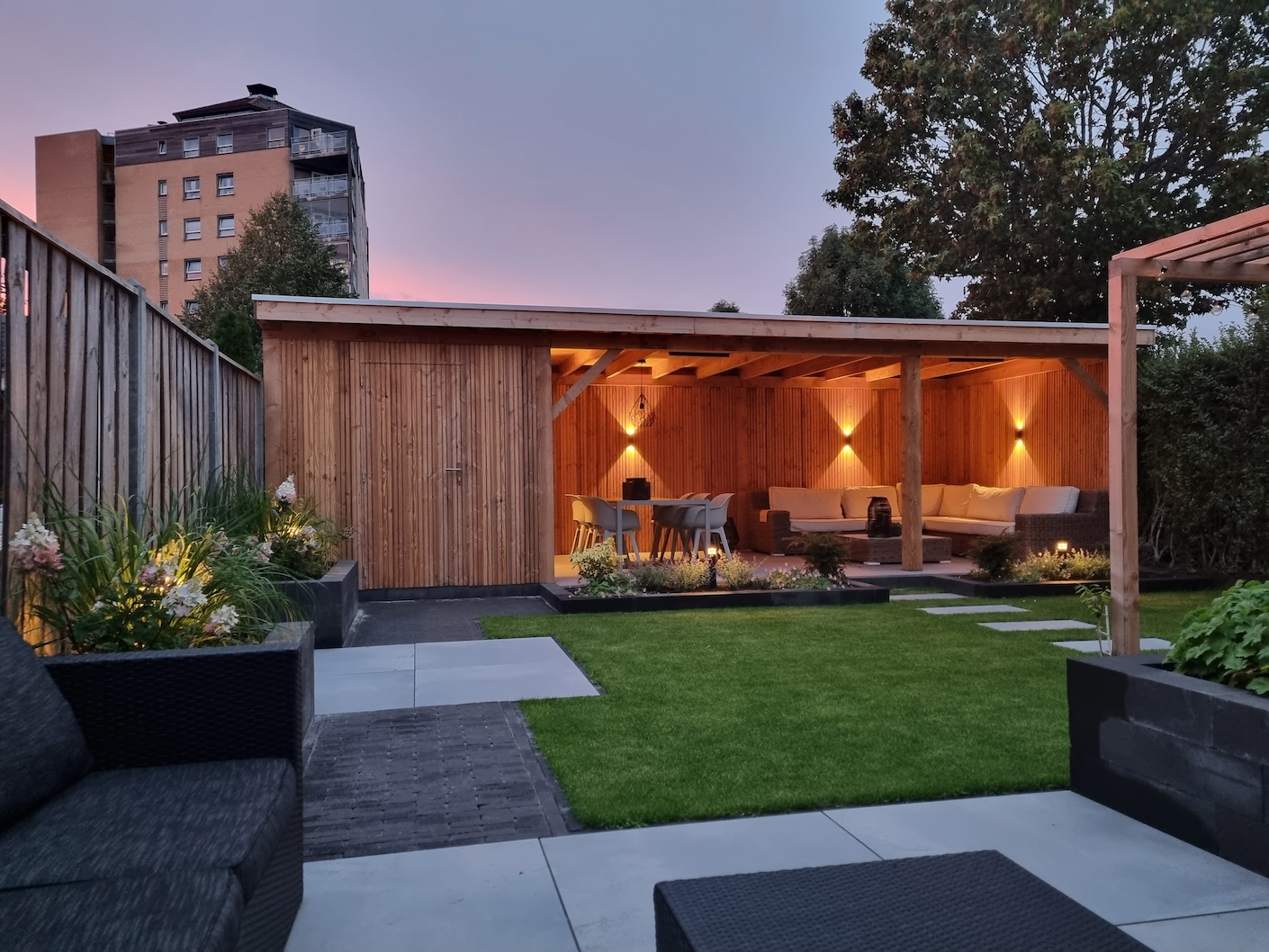 Een moderne tuin bij zonsondergang met een door Snijers Tuinen ontworpen en gebouwde houten pergola, sfeervolle verlichting, en comfortabele zitplaatsen onder een overkapping. Tuinrenovatie in Lelystad