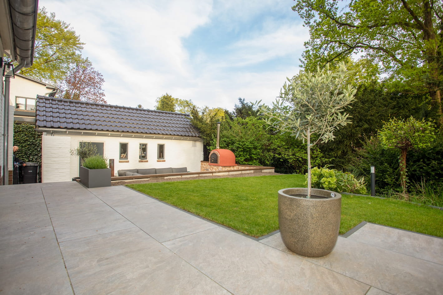 Tuin met betegeld terras, grasveld en een zitkuil met houtoven bij een tuinrenovatie in Hilversum door Snijers Tuinen.